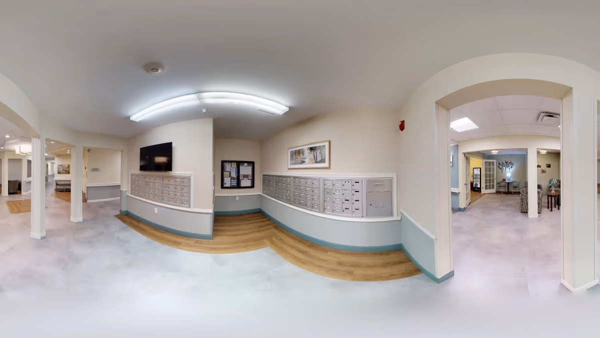 Interior view of a senior living facility hallway with multiple mailboxes mounted on the walls. The space has light-colored walls with teal trim, wood and tile flooring, and fluorescent ceiling lights. There is a flat-screen TV mounted above the mailboxes and a bulletin board with notices. The hallway leads to other rooms and common areas with seating visible in the background.