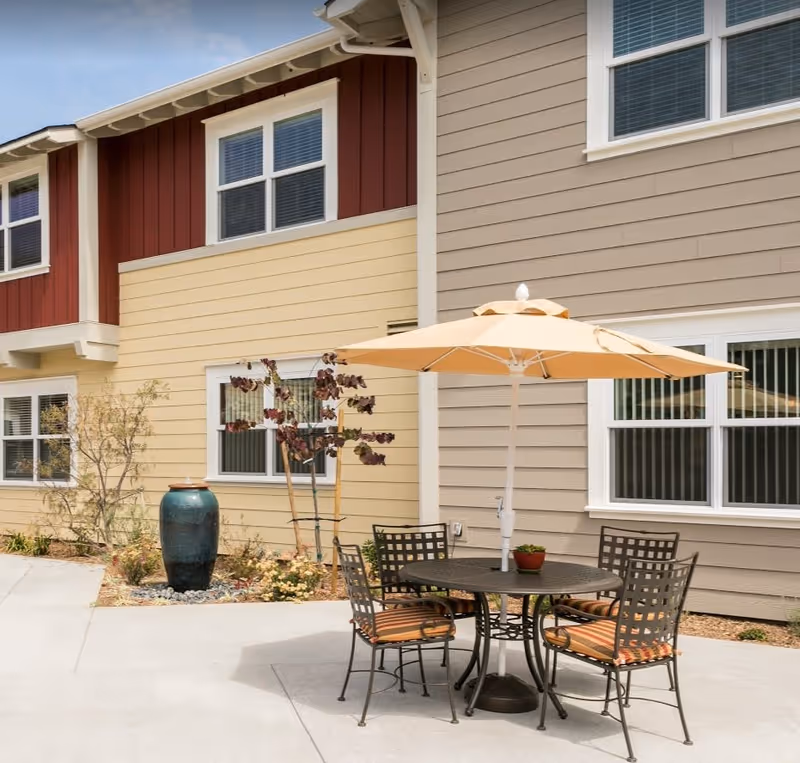 Outdoor patio area with a round metal table and four chairs with striped cushions under a large beige umbrella. The patio is adjacent to a building with beige, yellow, and red siding and several windows. There is a large decorative blue-green ceramic vase and some small plants near the building.