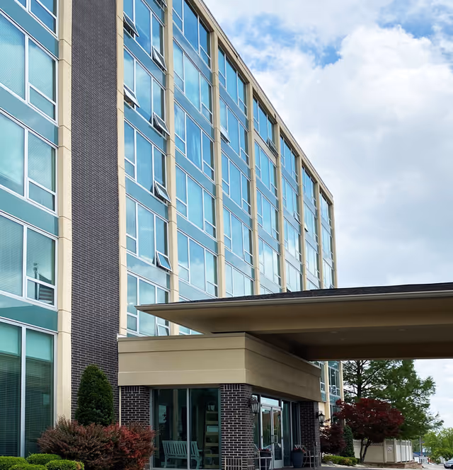 The front facade and covered entrance of a multi-story senior living building with large windows and landscaped shrubs.