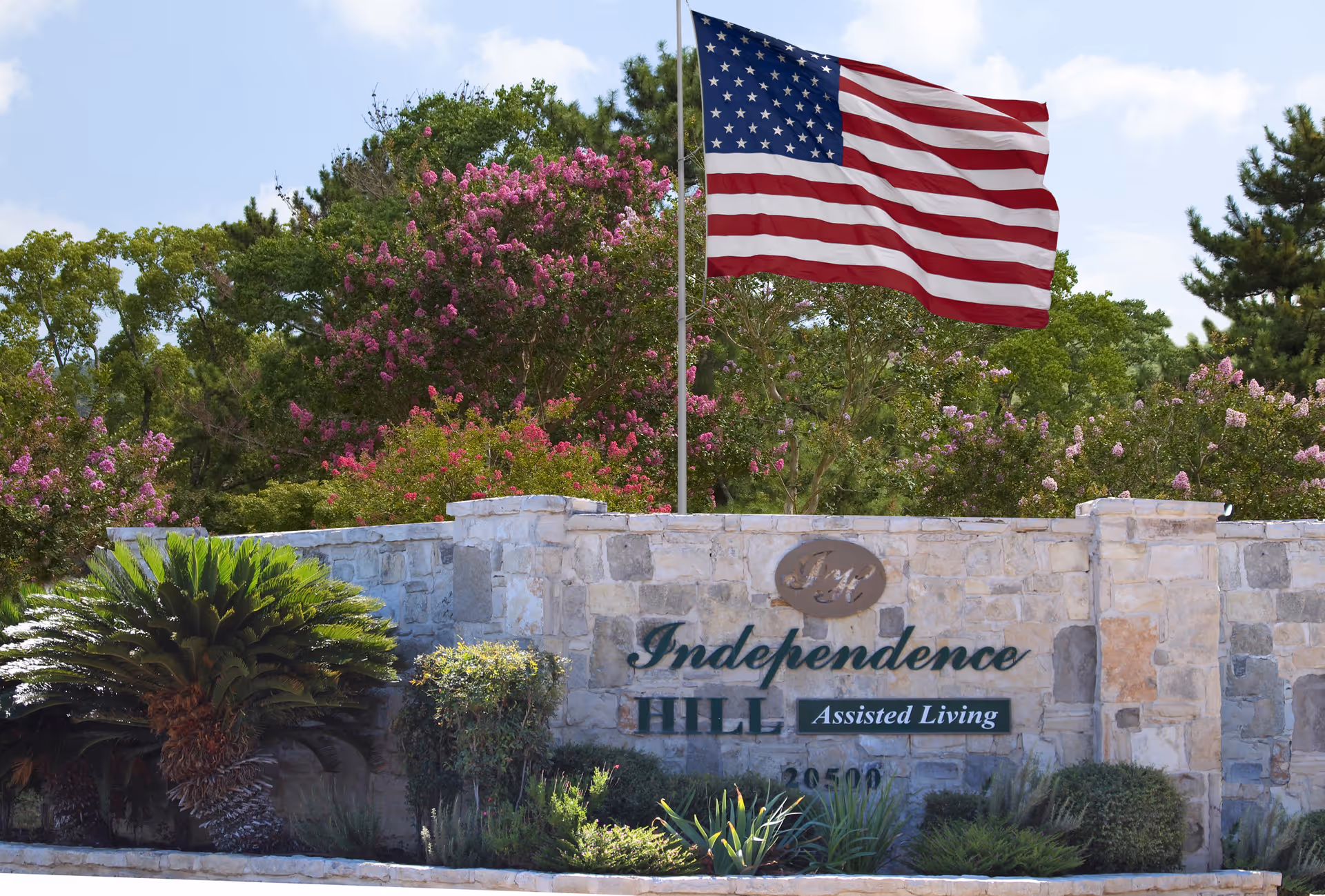 Stone entrance sign for Independence Hill Assisted Living with an American flag flying above it, surrounded by green shrubs and trees with pink flowers under a partly cloudy sky.