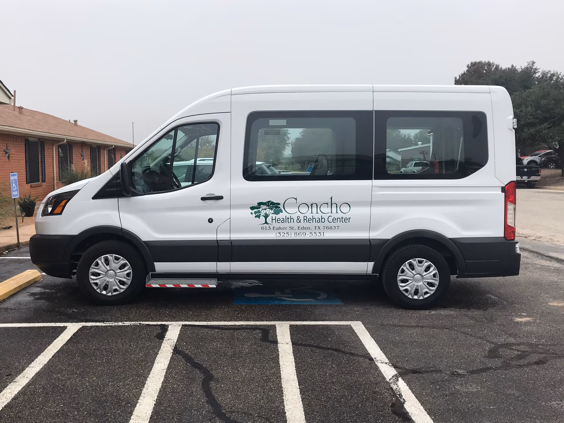 White Concho Health & Rehab Center passenger van parked in an outdoor lot beside a brick building.