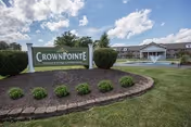 Landscaped entrance sign reading "CrownPointe" with the senior living facility and a fountain visible behind it under a partly cloudy sky.
