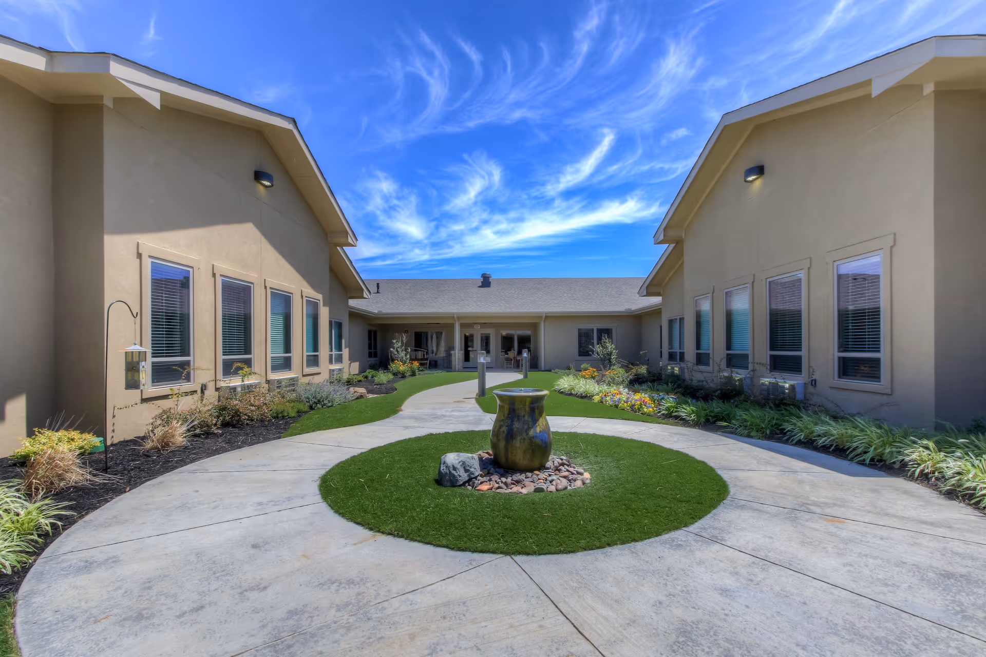 Outdoor courtyard area of Clear Fork Assisted Living and Memory Care with a circular concrete pathway surrounding a small grassy area featuring a decorative urn and rocks. The courtyard is bordered by beige buildings with multiple windows and landscaped with plants and flowers under a bright blue sky with wispy clouds.
