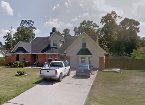 A residential house with a brick and white exterior, featuring a driveway with a white pickup truck and a blue sedan parked. Two people are standing near the vehicles. The house is surrounded by a lawn with some trees and bushes, under a partly cloudy sky.