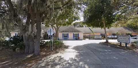 Entrance driveway leading to a single-story building with a sign that reads 'Royal Gardens of St Cloud' surrounded by trees and greenery.
