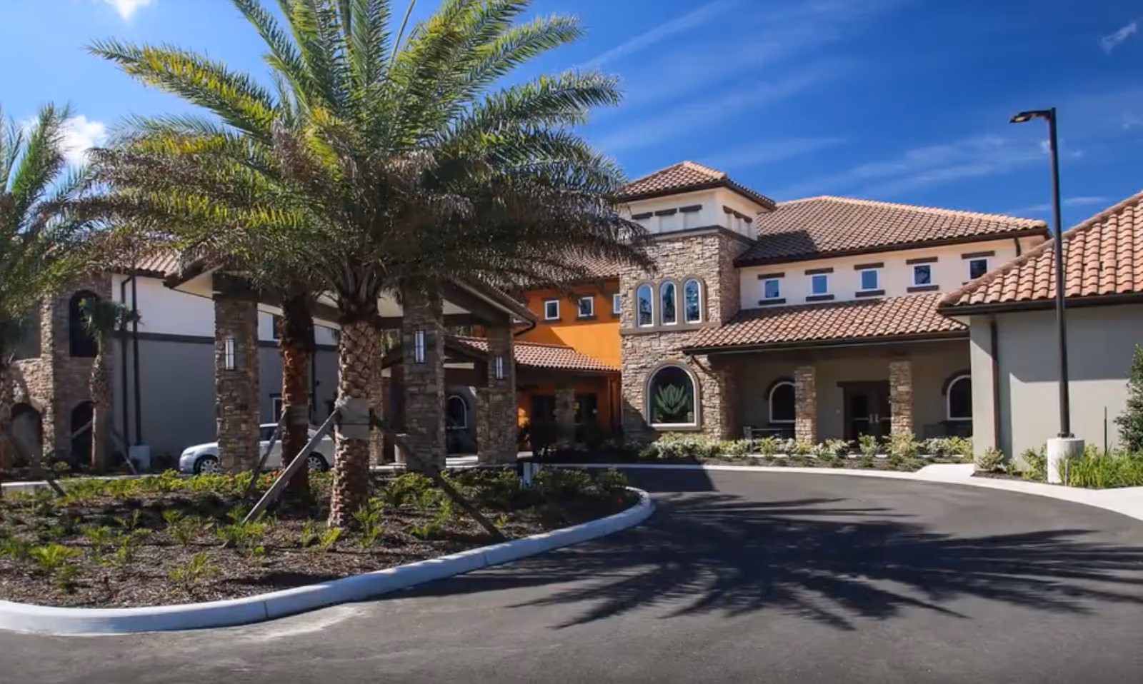 Exterior view of HarborChase of Palm Coast facility with a driveway, palm trees, and a building featuring stone and stucco walls under a clear blue sky.