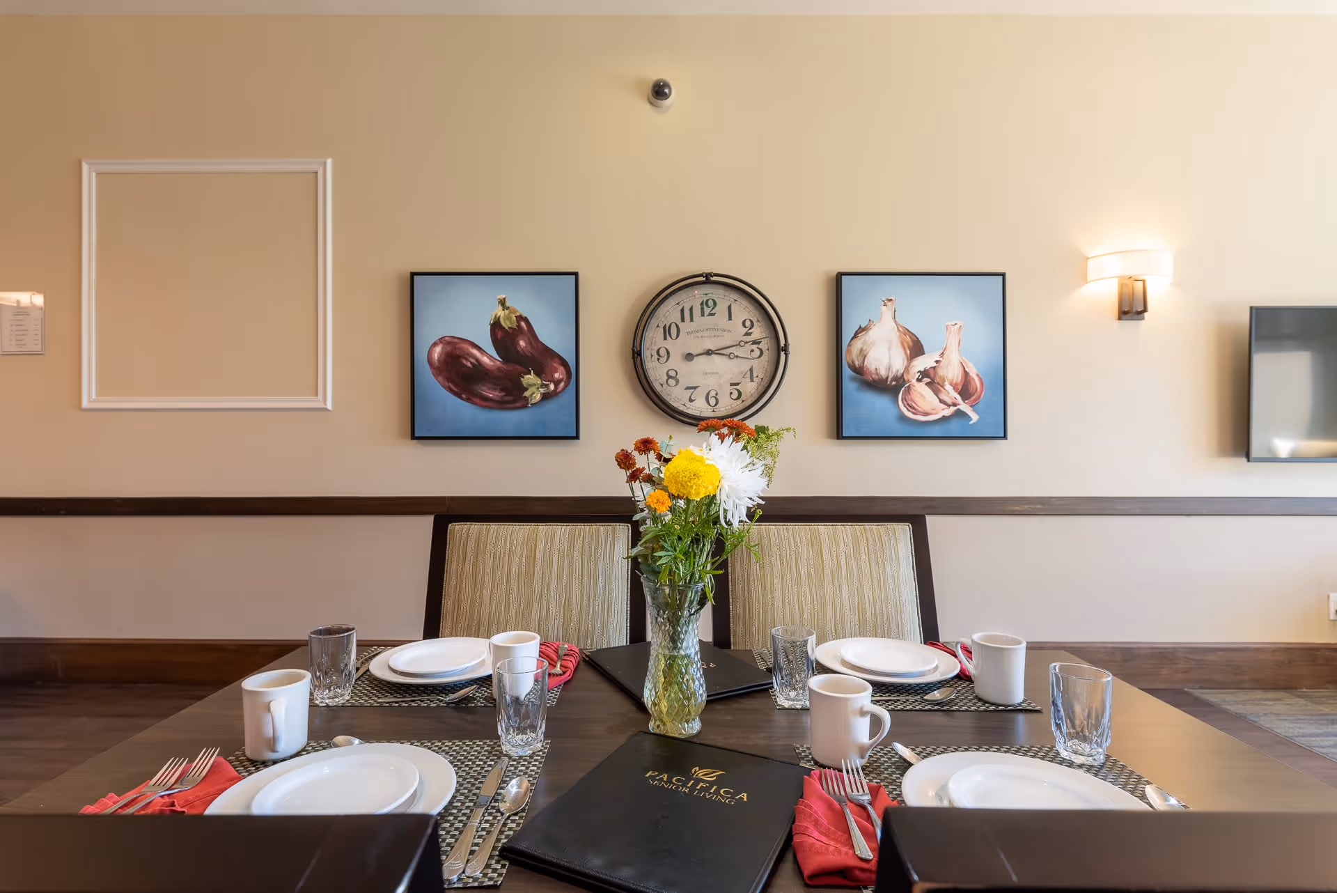 Table set for four with plates, mugs, glasses and a vase of flowers in a dining room with a wall clock and produce-themed artwork.
