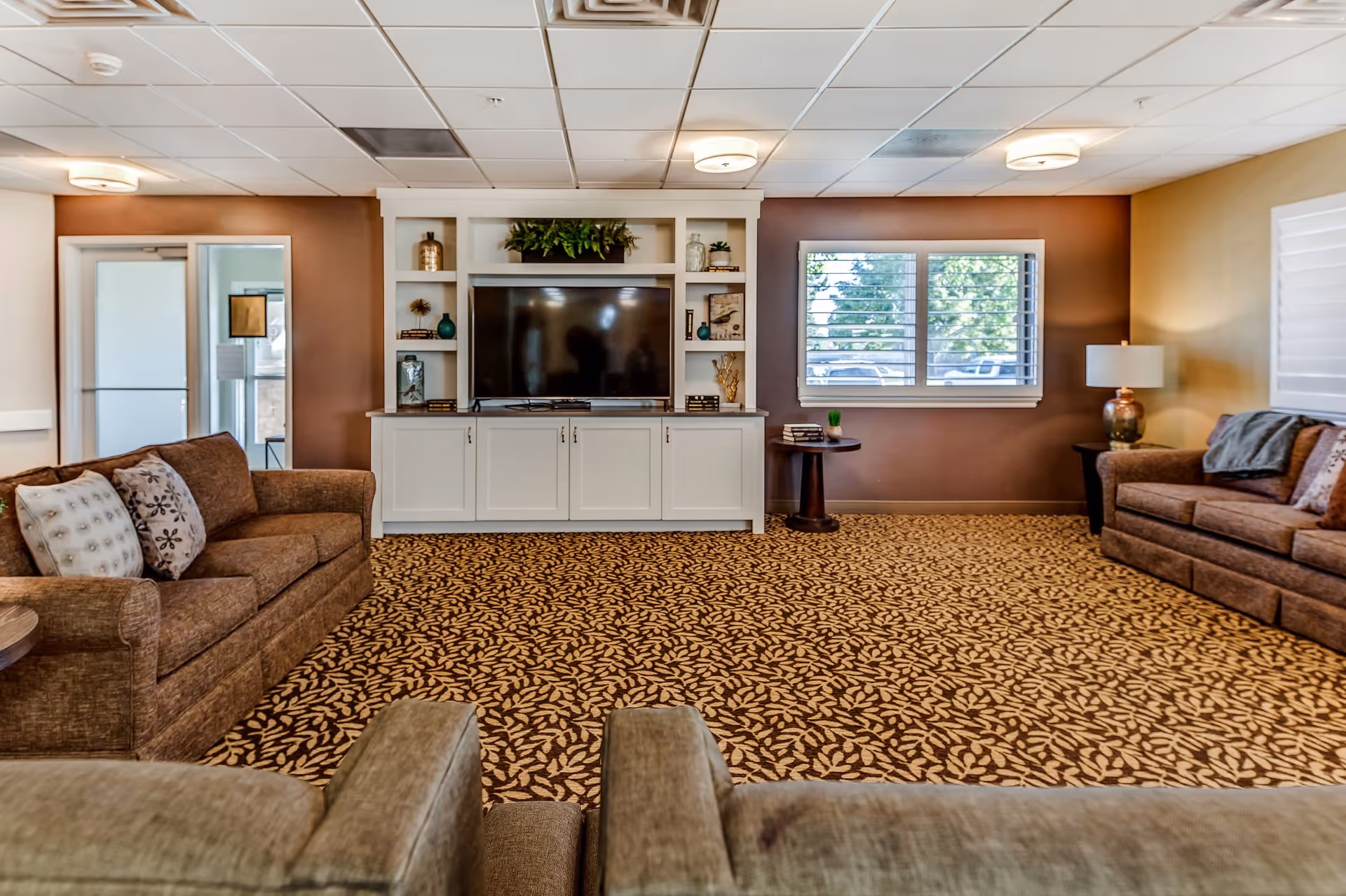 A cozy living room with patterned carpet, two brown sofas with decorative pillows, a large flat-screen TV mounted on a white built-in cabinet with shelves displaying decorative items, a window with white blinds, and a table lamp on a side table.