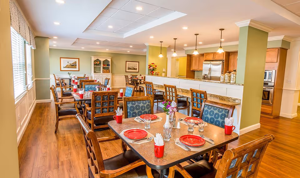 Bright communal dining room with multiple set tables and wooden chairs and a serving counter/kitchen area in the background.