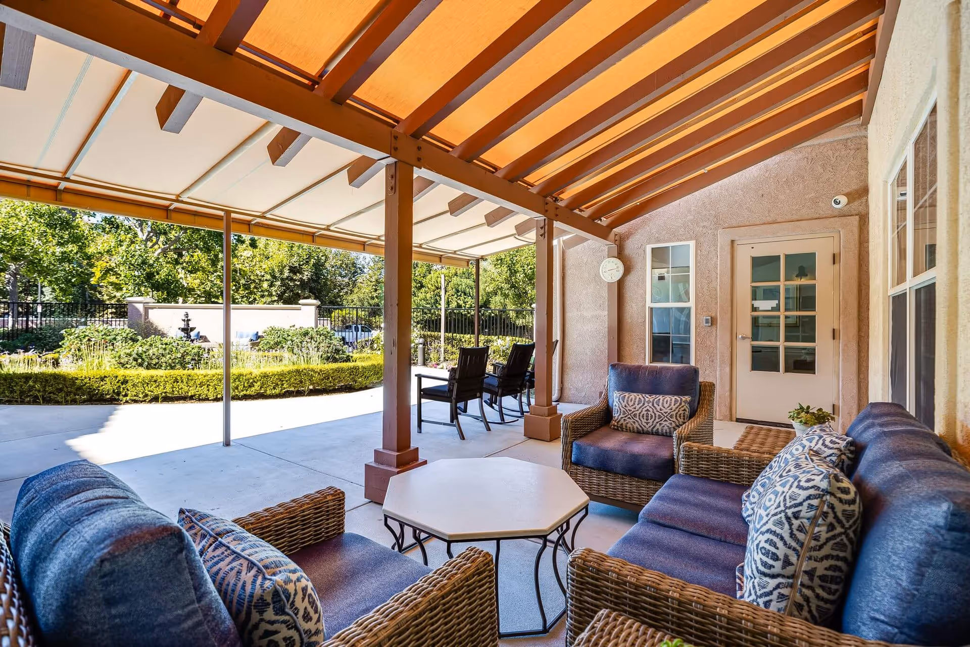 Covered outdoor patio with wicker seating, patterned cushions, a central table, and a view of a landscaped garden.