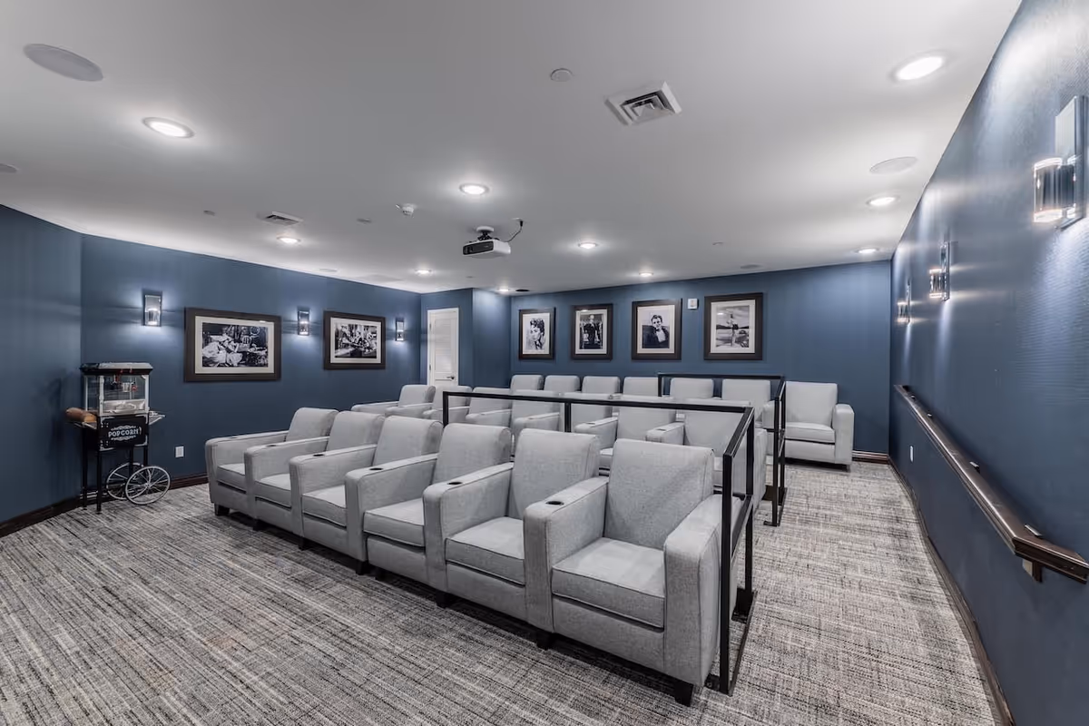 A small theater room with two rows of gray upholstered armchairs separated by black metal railings. The walls are painted blue and decorated with black and white framed photographs. There is a popcorn machine in the corner and ceiling lights illuminating the room.