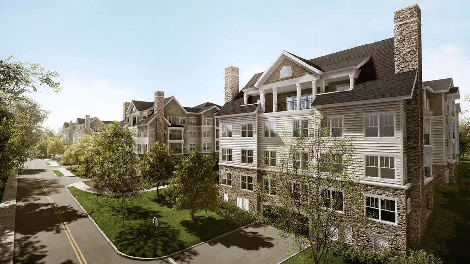 Exterior view of a multi-story senior living facility building with stone and siding facade, surrounded by trees and greenery along a paved road under a clear sky.