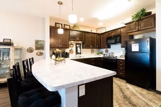 Modern kitchen with dark wood cabinets, black refrigerator, and a curved white countertop island with black cushioned bar stools. The kitchen is well-lit with pendant lights and recessed ceiling lights. There are decorative plants and framed pictures on the walls and cabinets.