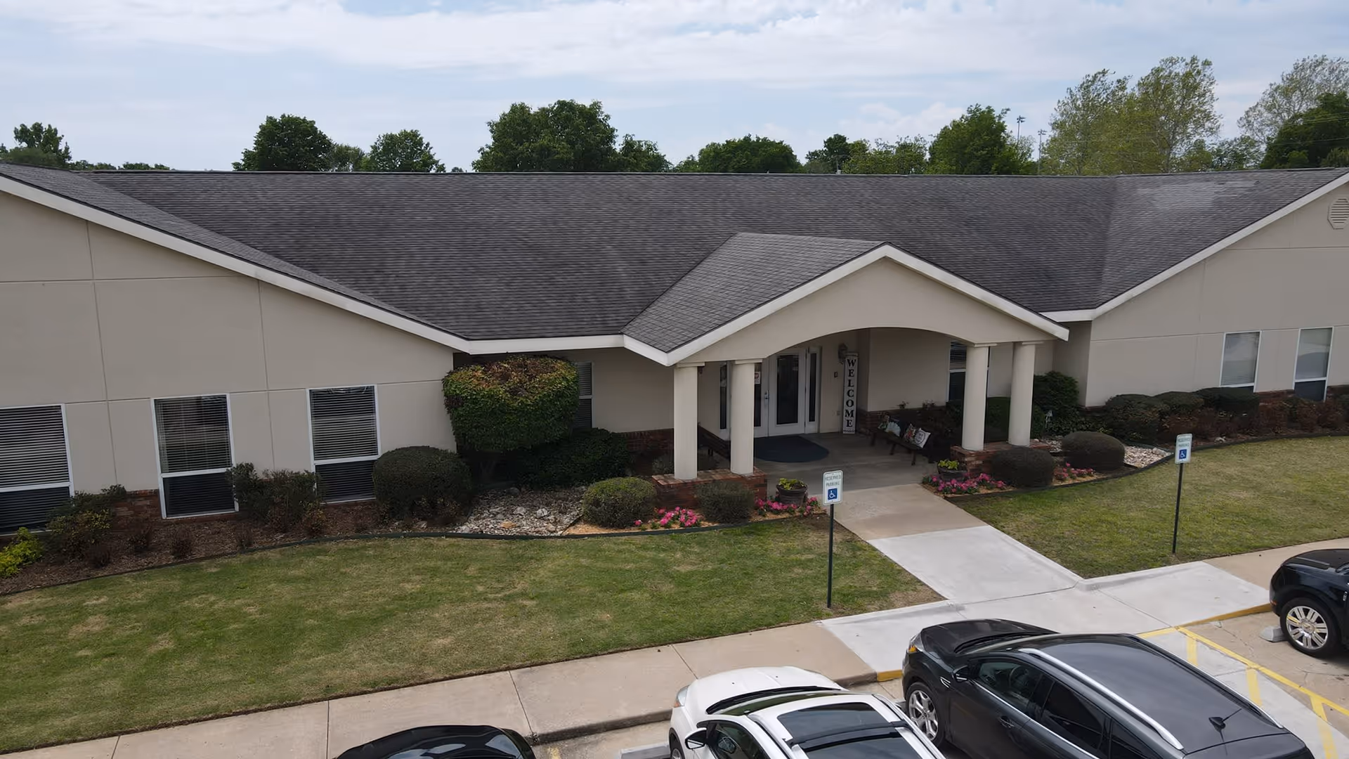 Exterior view of a single-story assisted living facility building with a covered entrance supported by columns. There is a walkway leading to the entrance, surrounded by well-maintained landscaping including bushes and flowers. Several cars are parked in front of the building in designated parking spaces.