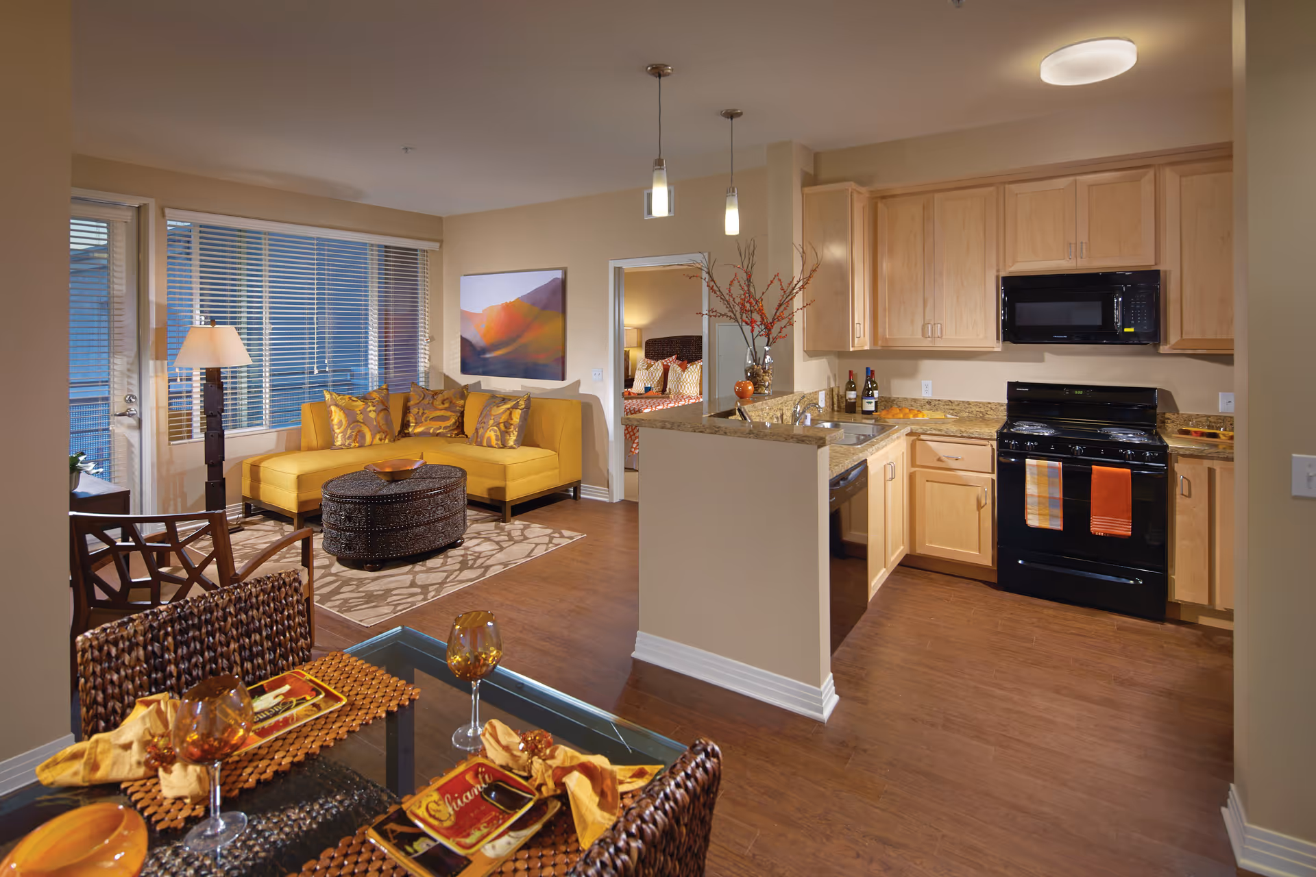 Interior view of a senior living apartment featuring an open kitchen with light wood cabinets, black appliances, and granite countertops. Adjacent to the kitchen is a living area with a mustard yellow sectional sofa, patterned pillows, a round dark coffee table, and a floor lamp near large windows with blinds. In the foreground, there is a dining area with a glass table set with placemats, napkins, and amber-colored glasses. A doorway leads to a bedroom with a bed visible in the background.