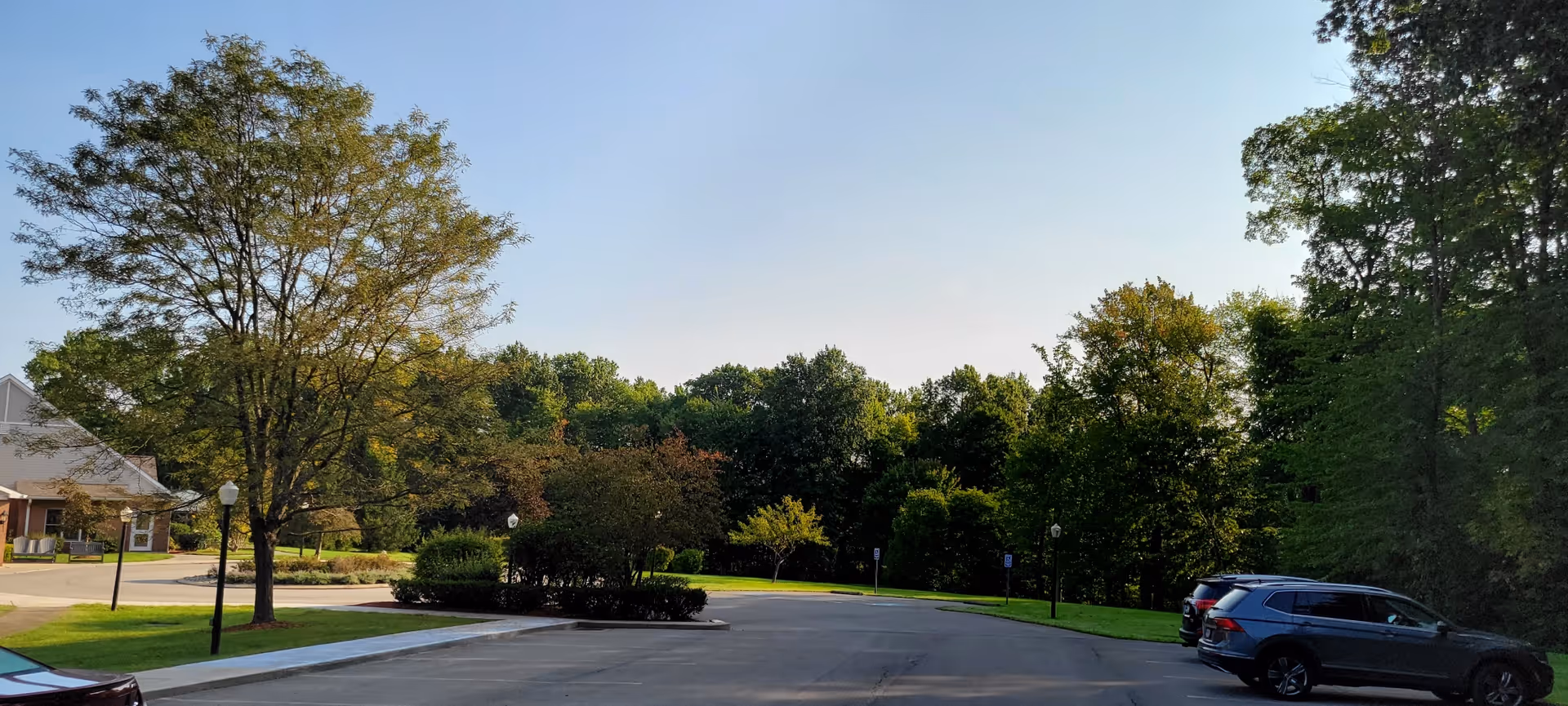 Parking lot area with several parked cars, surrounded by green trees and bushes under a clear blue sky. A building is partially visible on the left side of the image.