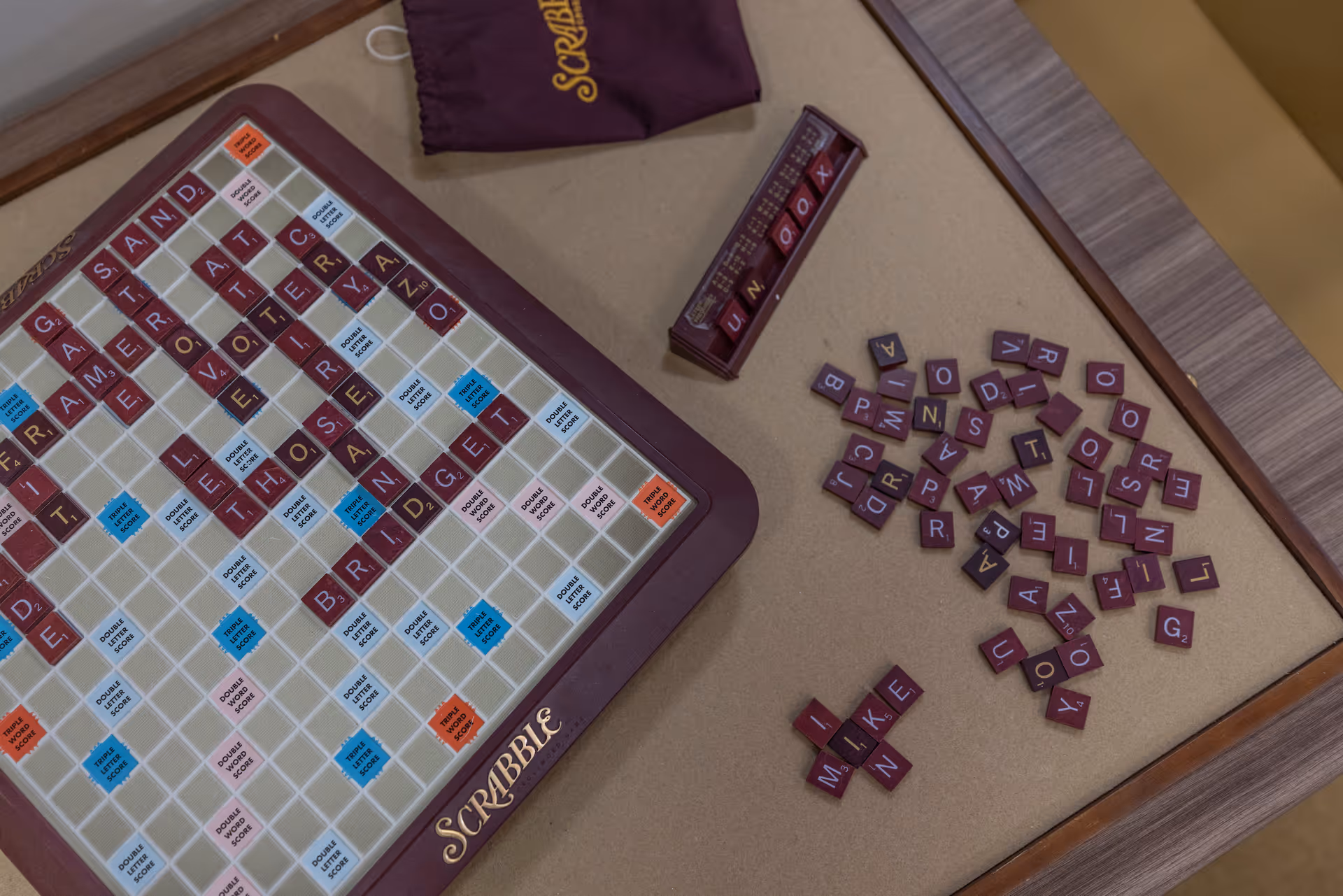 A Scrabble board game in progress on a wooden table with letter tiles scattered around and some tiles placed on the board forming words like 'BRING' and 'THOSE'. A purple Scrabble tile holder and a purple Scrabble bag are also visible.