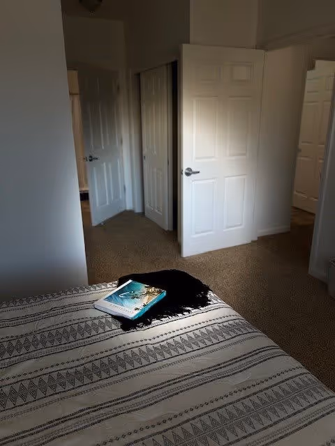 View of a bedroom corner with a bed covered in a patterned blanket, a book and a black cloth on the bed. The room has beige carpet and white walls with multiple white doors leading to other rooms or closets.