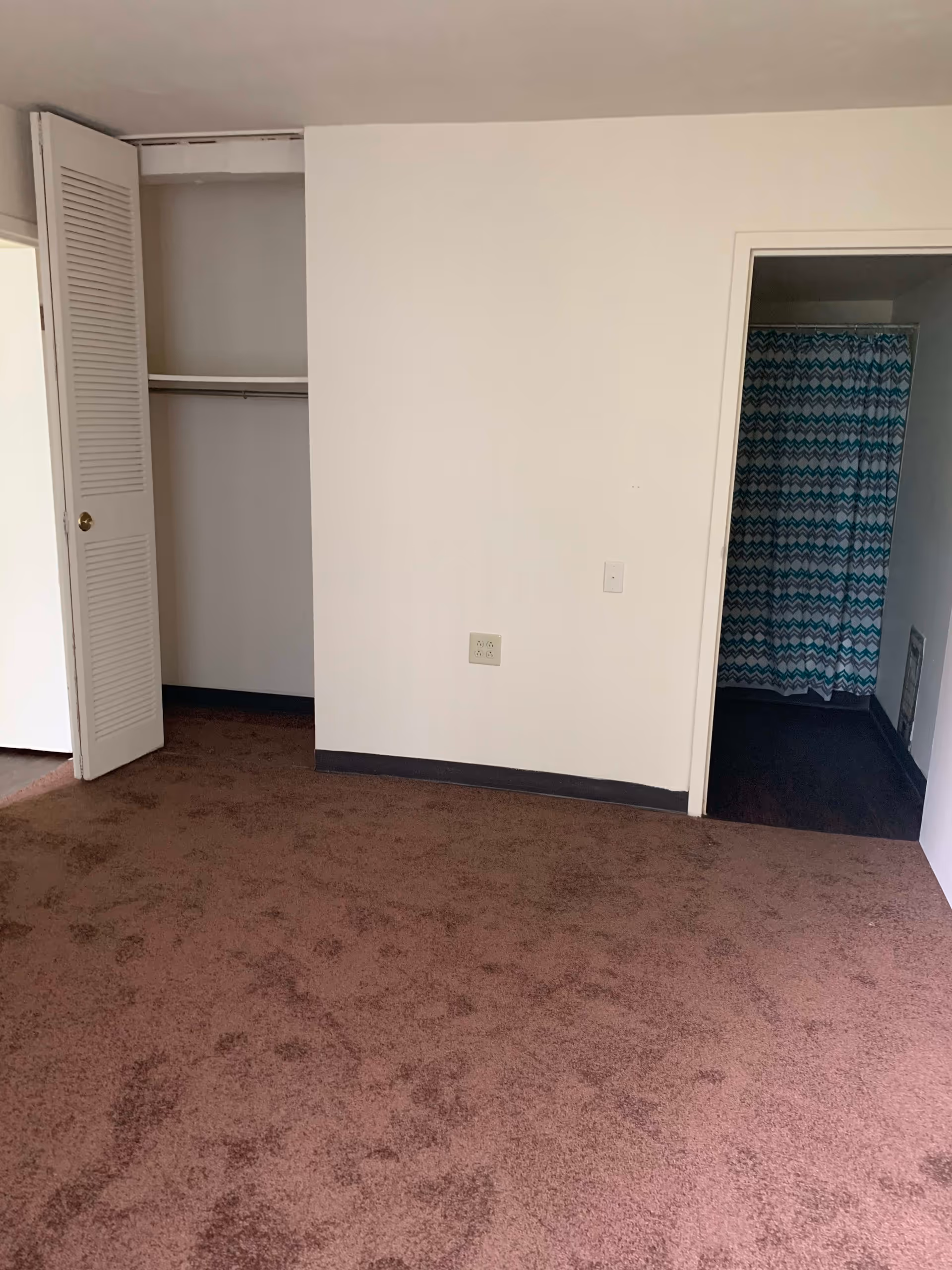 Empty room with brown carpet, an open closet with white louvered doors on the left, and a doorway on the right leading to a bathroom with a blue and white patterned shower curtain.