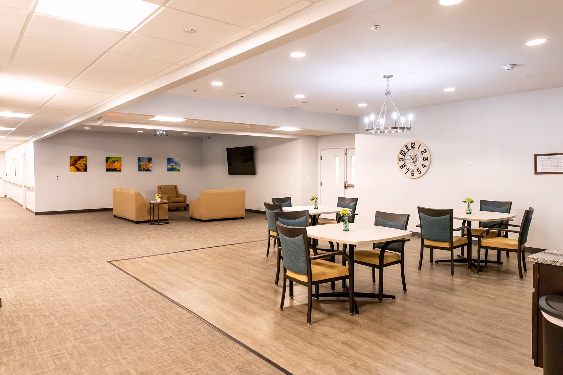 A spacious senior living facility common area with several tables and chairs arranged for dining or socializing. The room features a modern chandelier, a large wall clock, and a seating area with beige sofas and a wall-mounted TV. The walls are decorated with colorful artwork, and the floor transitions from carpet to wood-like flooring.