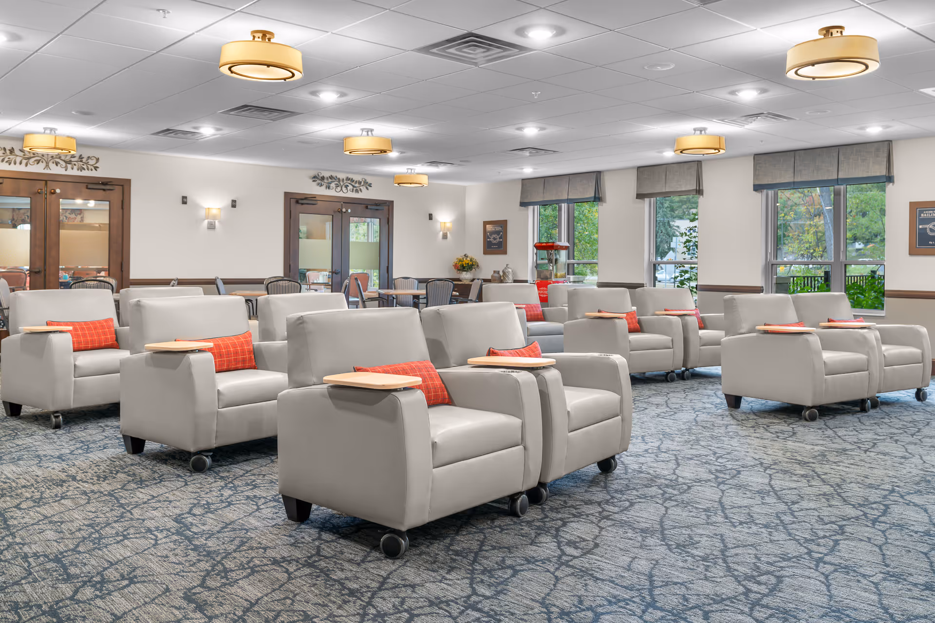 Spacious senior living common room with rows of light-gray upholstered recliners on wheels, red accent pillows and small attached trays beneath ceiling lights and windows.