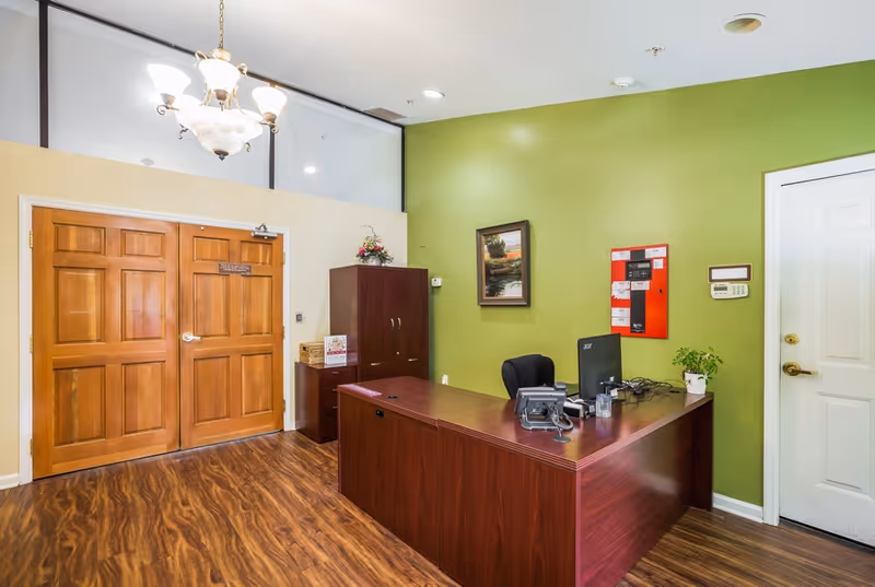 Reception area with a wooden desk, office chair, computer, telephone, and a small plant. The walls are painted green and beige, with a framed picture and a red fire alarm panel on the green wall. There are double wooden doors on the left and a white door on the right. A chandelier hangs from the ceiling, and the floor has a wood-like finish.