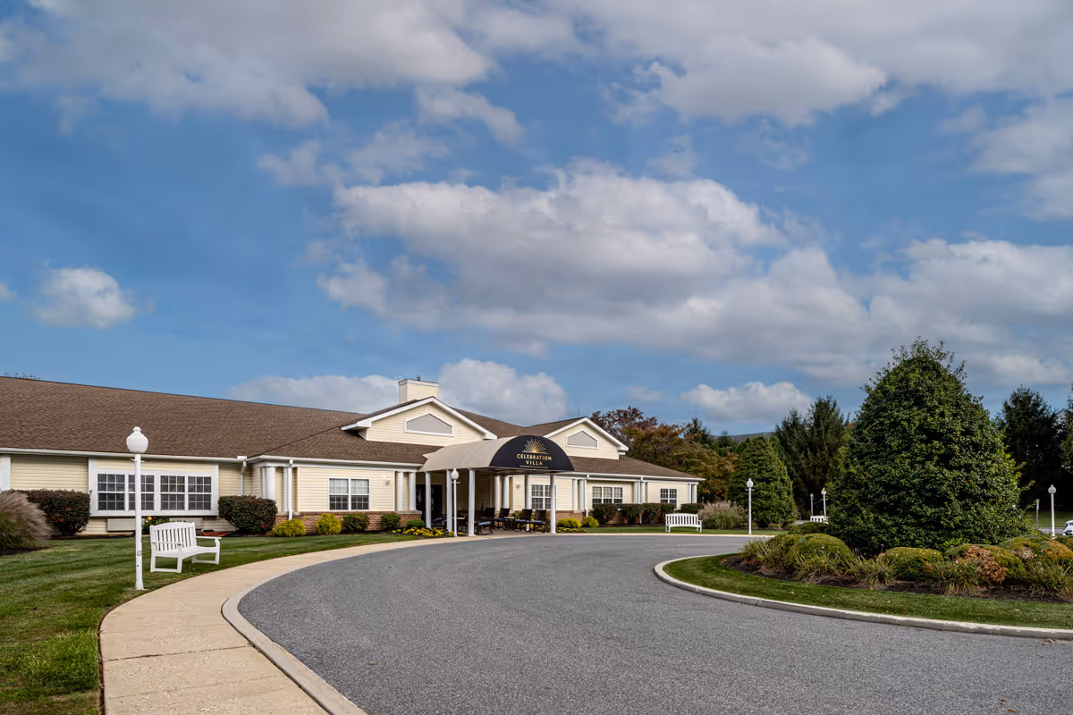Exterior view of Celebration Villa of York, a single-story building with a brown roof and light yellow siding. The entrance has a covered canopy with the facility's name on it. There is a curved driveway leading to the entrance, surrounded by well-maintained landscaping including bushes, trees, and grass. White benches and lamp posts line the walkway near the building. The sky is partly cloudy with blue patches.