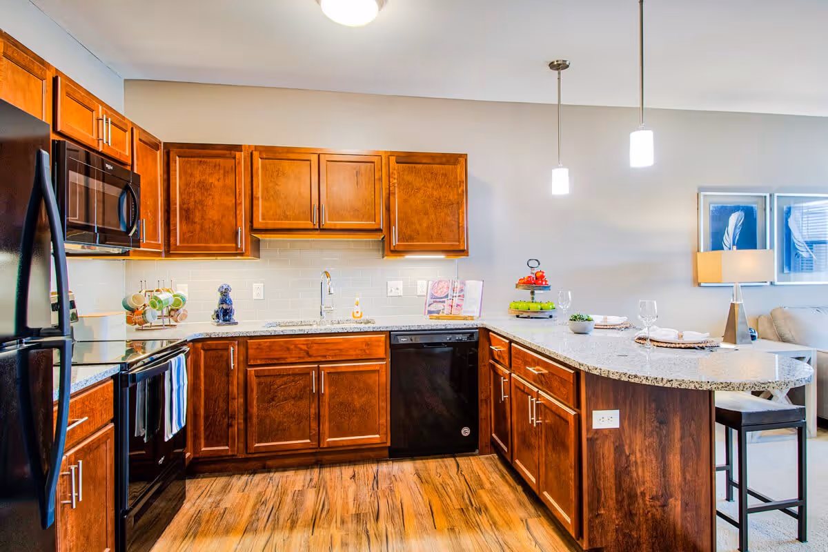 Modern kitchen with wooden cabinets, black appliances including refrigerator, microwave, stove, and dishwasher, granite countertops, a kitchen island with bar stools, pendant lights, and a view into a living area with a lamp and framed artwork.