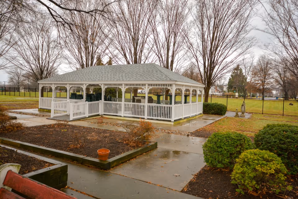 A white gazebo with a gray roof situated in a garden area with leafless trees and green bushes. The ground is wet, indicating recent rain, and there are paved walkways surrounding the gazebo. In the background, there is a black metal fence enclosing the area.