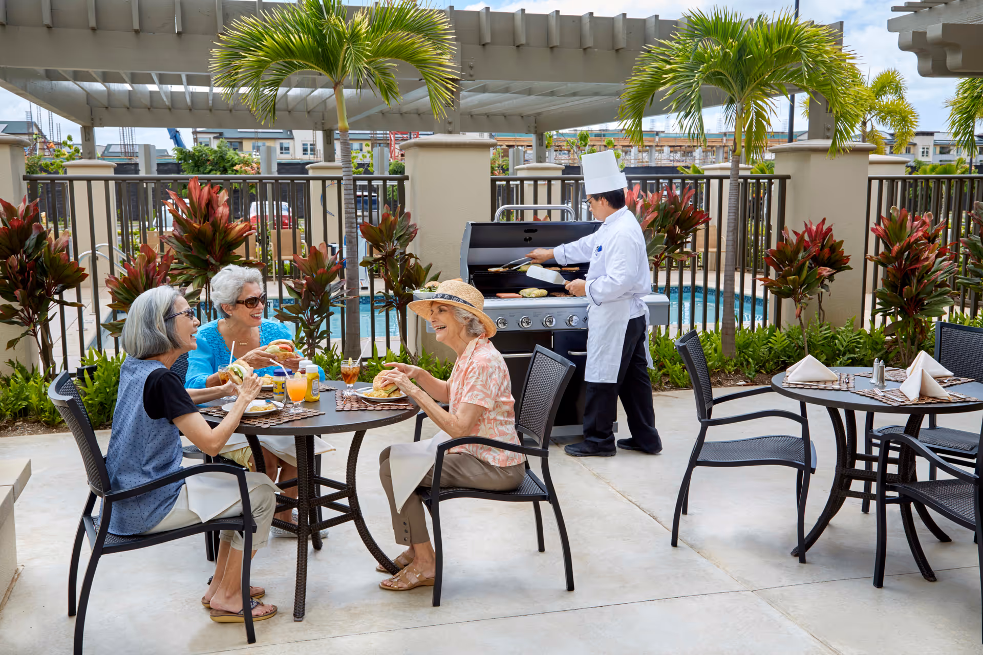 Three elderly women sitting around a round outdoor table enjoying a meal and drinks while a chef in a white uniform and hat grills food on a barbecue grill nearby. The setting is a patio area with palm trees, plants, and a swimming pool in the background under a pergola.