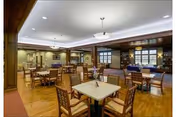 Dining room with multiple wooden tables and cushioned chairs under pendant lights in a warm, wood-paneled interior with large windows.