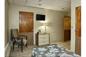 A simple bedroom with a bed covered in a floral patterned bedspread, a wooden door, a small dresser with a lamp and picture frame on top, a wall-mounted TV, and a cushioned chair with armrests.