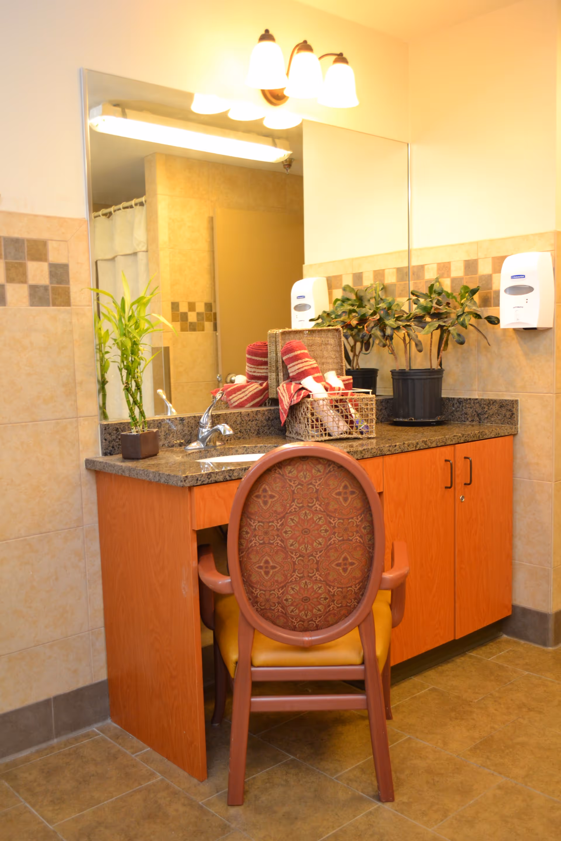 A bathroom vanity area with a granite countertop, a sink, and a large mirror. On the countertop, there are two potted plants and a basket containing rolled red and white towels. A decorative chair with a patterned backrest and yellow seat is placed in front of the vanity. The walls are tiled with beige and brown tiles, and there is a soap dispenser mounted on the wall.