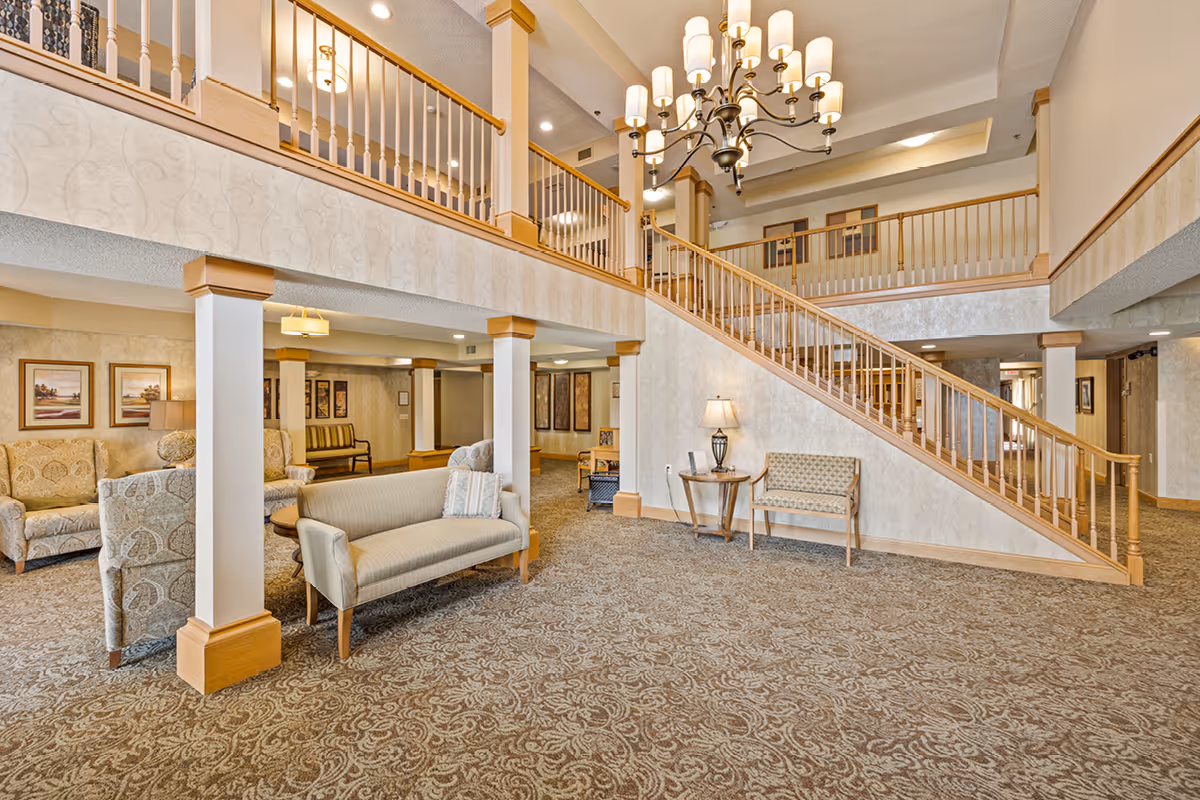Spacious senior living facility common area with patterned carpet, beige walls, and wooden accents. The room features a staircase with wooden railings leading to an upper floor balcony. There are several seating arrangements including sofas, armchairs, and benches, along with framed artwork on the walls and a chandelier hanging from the ceiling.
