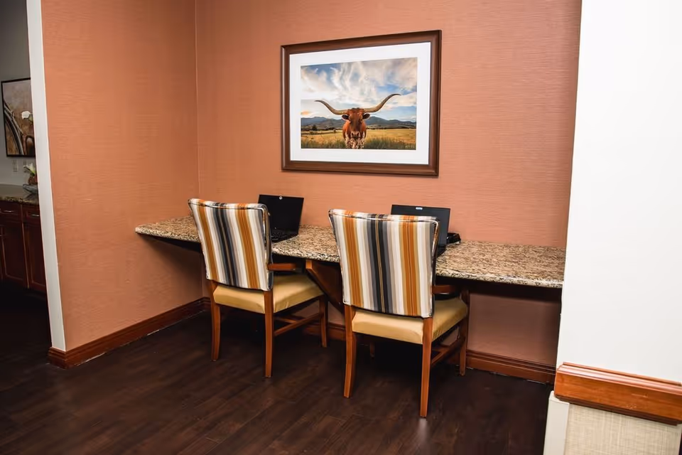 A small workspace area with a granite countertop attached to the wall, two striped upholstered chairs, and two laptops placed on the countertop. A framed picture of a longhorn cow in a field hangs on the orange-brown wall above the workspace. The floor is dark wood, and part of a kitchen or cabinet area is visible to the left.