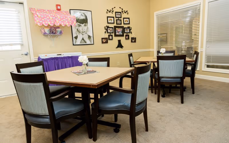 A small dining area with two square tables, each surrounded by four chairs with dark wooden frames and light blue upholstery. The tables have small flower vases with white flowers and some magazines. The walls are beige with framed pictures, including a large black and white portrait of a woman. There is a window with blinds and a door with a window on the left side. A purple skirted table with a pink and white decorative canopy is against the wall.