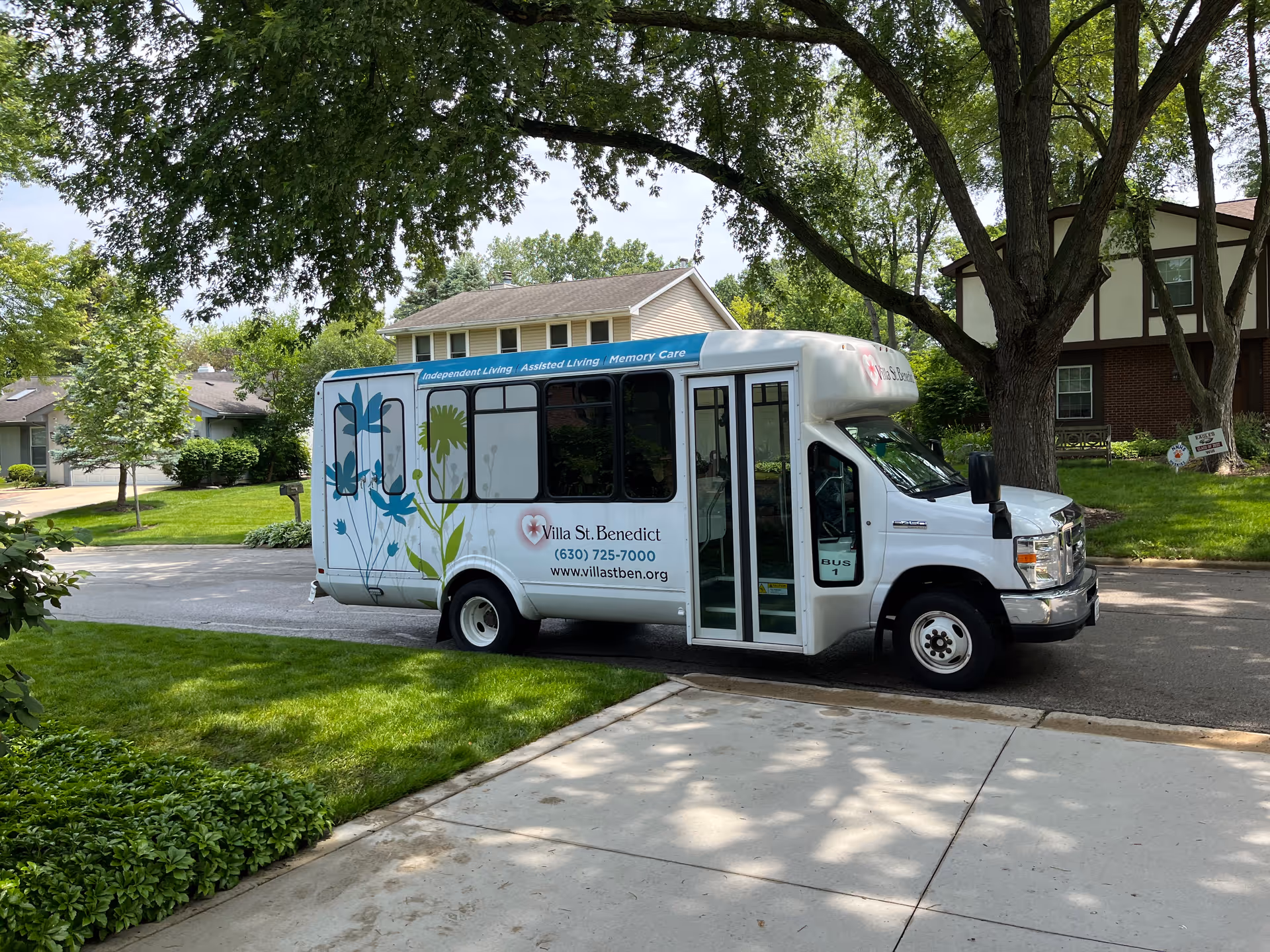 A white shuttle bus parked on a residential street near green lawns and trees. The bus has floral graphics and text on the side that reads 'Villa St. Benedict' with a phone number and website. The bus is labeled for Independent Living, Assisted Living, and Memory Care services.