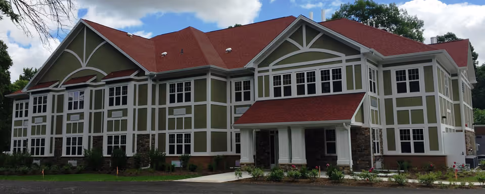 Large two-story senior living building with green and white siding, a red roof, many windows, and a covered front entrance.