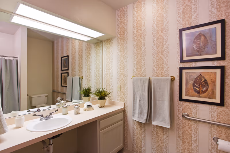 Well-lit bathroom with a sink and vanity, large mirror, patterned wallpaper, towel rack, framed leaf artwork, and a toilet and shower in the background.