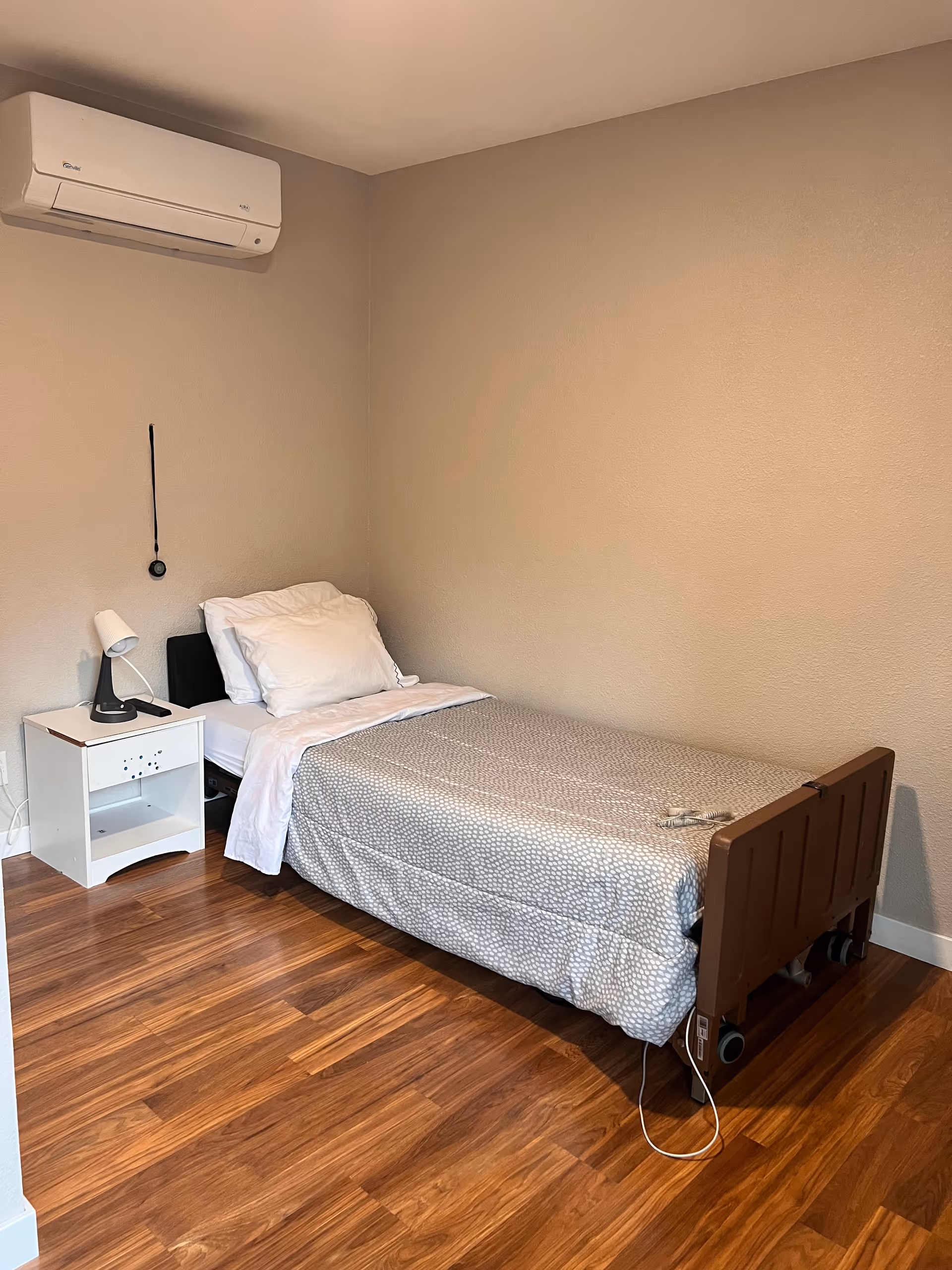 Small single-occupancy bedroom with an adjustable single bed, nightstand and lamp, wall-mounted air conditioner, and wood flooring.