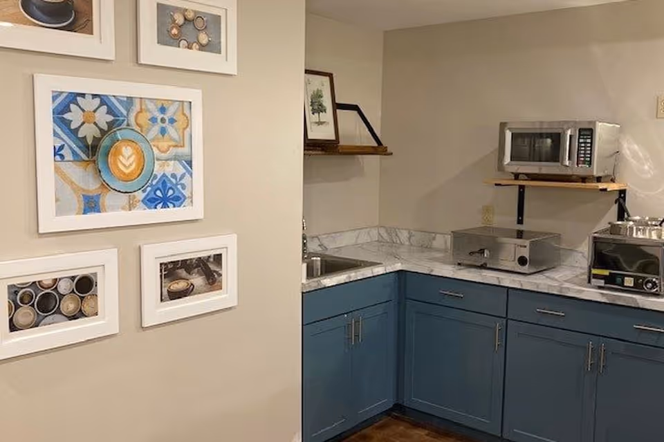 A small kitchen area with blue lower cabinets and white marble countertops. On the countertop, there is a microwave, a toaster oven, and a small oven. Above the countertop, there is a wooden shelf with a framed picture. On the adjacent wall, there are five framed pictures featuring coffee cups and coffee-related images.