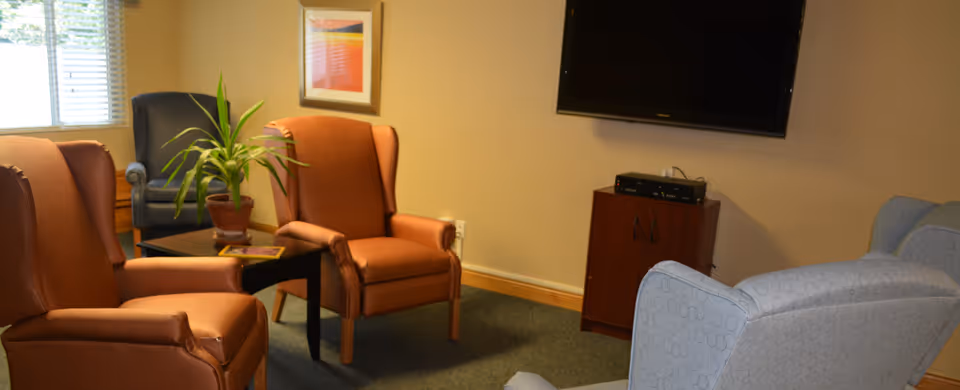 A cozy living room area with several armchairs arranged around a small black coffee table with a potted plant on it. There is a window with blinds letting in natural light, a framed picture on the wall, and a flat-screen TV mounted above a wooden cabinet.