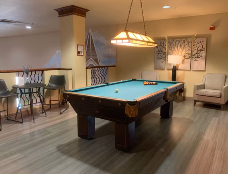 A well-lit recreational room featuring a pool table with balls arranged for a game. The room has light wood flooring, a hanging rectangular light fixture above the pool table, two beige armchairs with a lamp and tree artwork on the wall behind them, and a small round table with two chairs near a railing with decorative balusters.