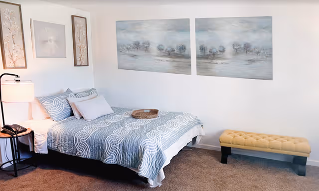 A neatly made bed with a blue and white patterned comforter and pillows in a senior living bedroom. There is a small round side table with a lamp and a phone next to the bed. Above the bed are three framed artworks, and on the adjacent wall are two large landscape paintings. A beige tufted bench is placed against the wall on the right side of the room. The floor is carpeted and the walls are painted white.