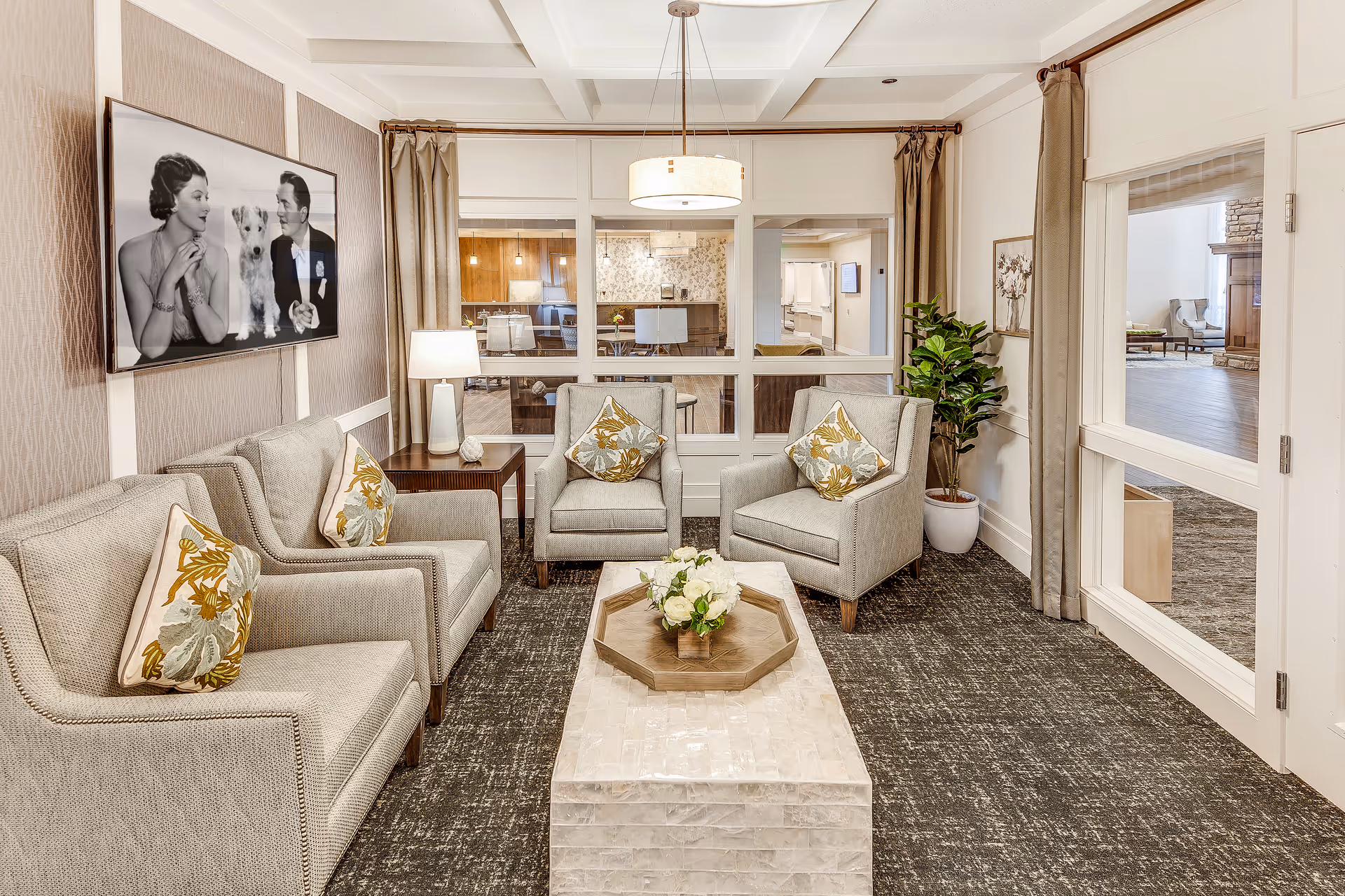 A cozy living room with four beige armchairs, each with a floral cushion, arranged around a rectangular coffee table with a flower centerpiece. A side table with a lamp is next to one armchair. The room has large windows looking into another room, beige curtains, a patterned carpet, and a black and white photo of a man, woman, and dog on the wall.