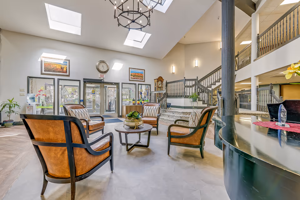 Bright senior living lobby with leather chairs arranged around a coffee table, glass entrance doors, and a staircase leading to an upper balcony.