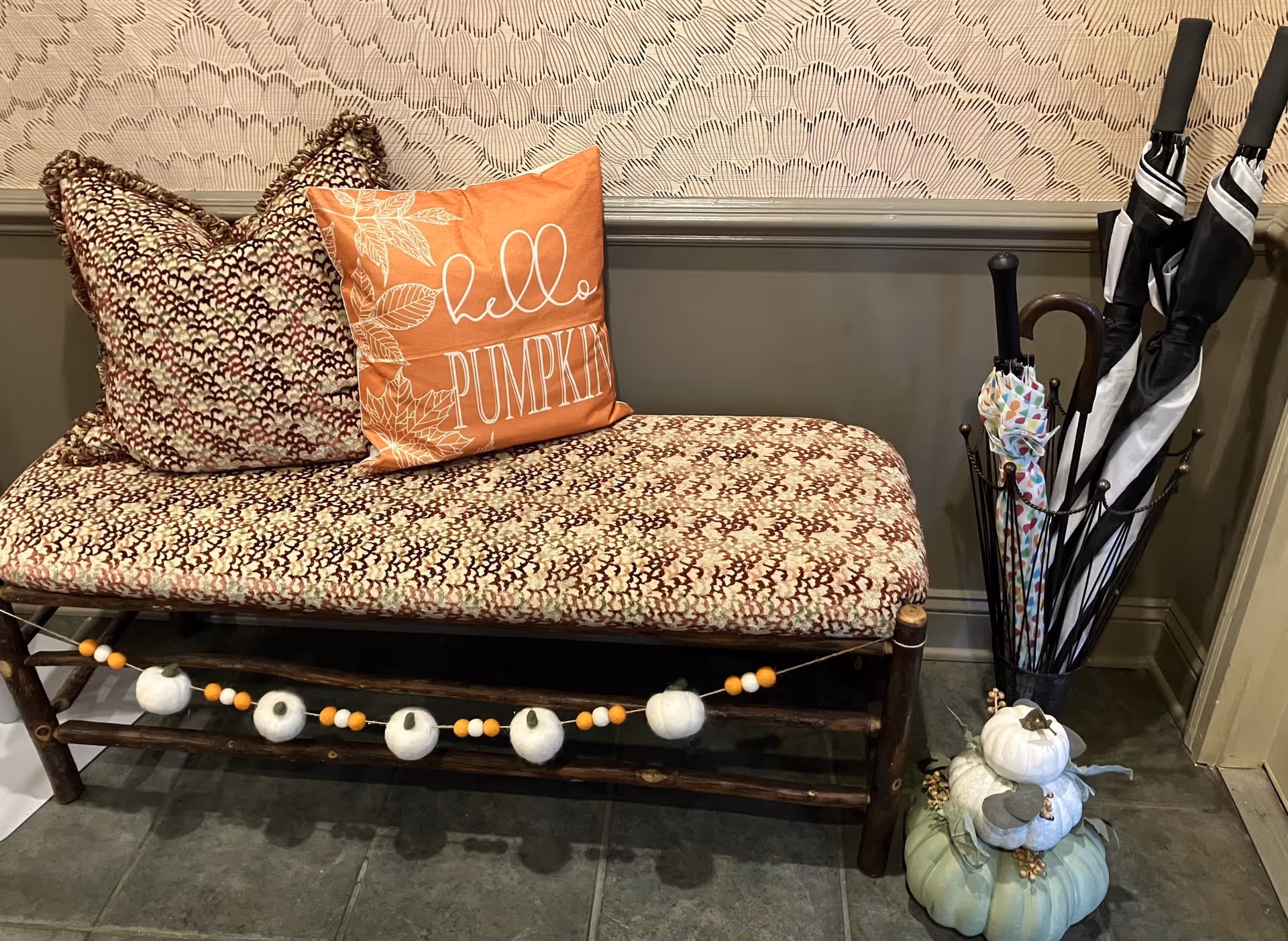 A cushioned bench with two patterned pillows, one of which is orange with the text 'hello PUMPKIN' written on it. The bench has a decorative garland with white and orange felt balls and pumpkins hanging on the front. Next to the bench is an umbrella stand holding several umbrellas and a decorative arrangement of pumpkins on the floor. The wall behind has a textured wallpaper and a chair rail molding.