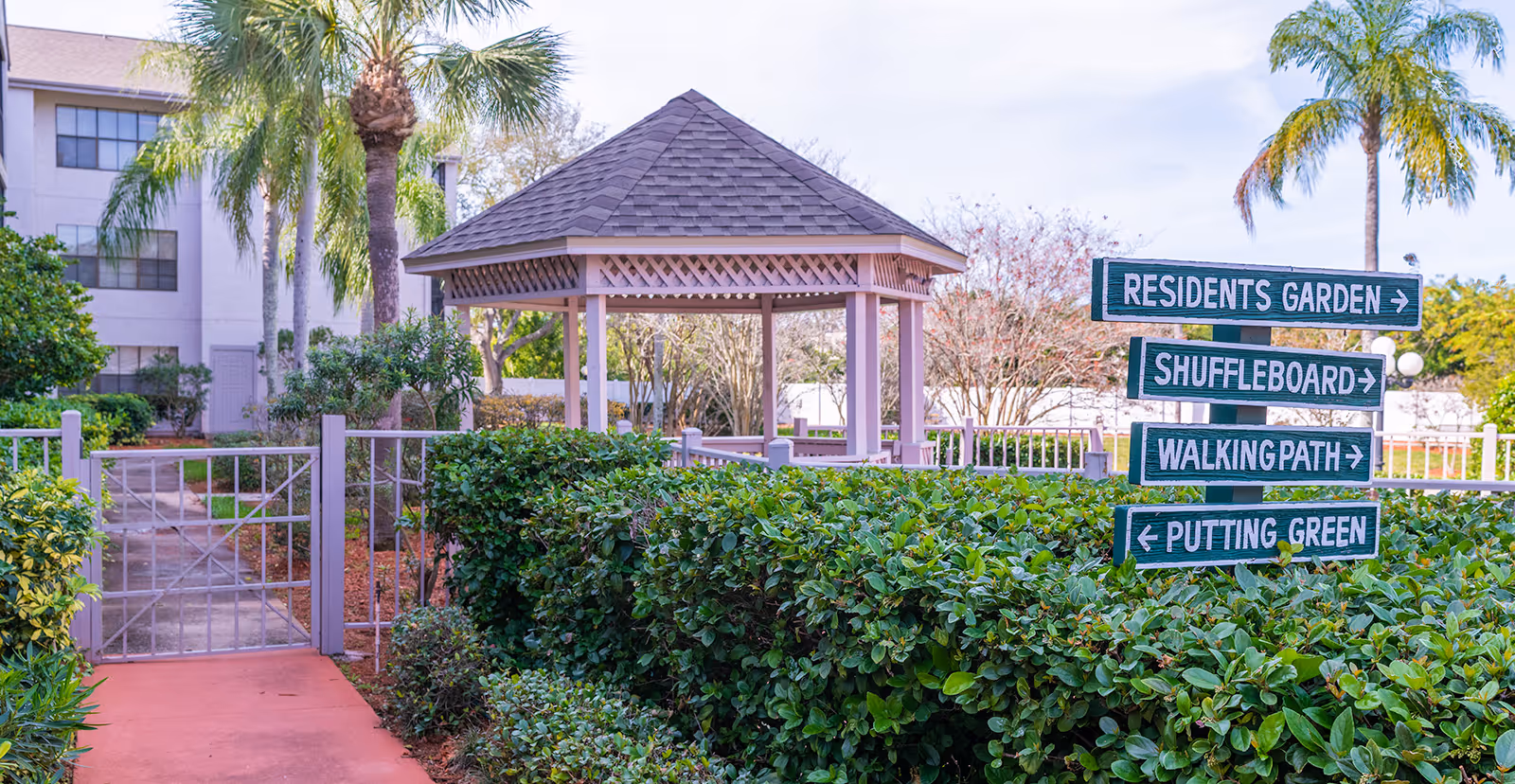 Outdoor area of Regency Residence - Sky Active Living featuring a gazebo surrounded by greenery and palm trees. A signpost indicates directions to the Residents Garden, Shuffleboard, Walking Path, and Putting Green. A building is visible in the background.
