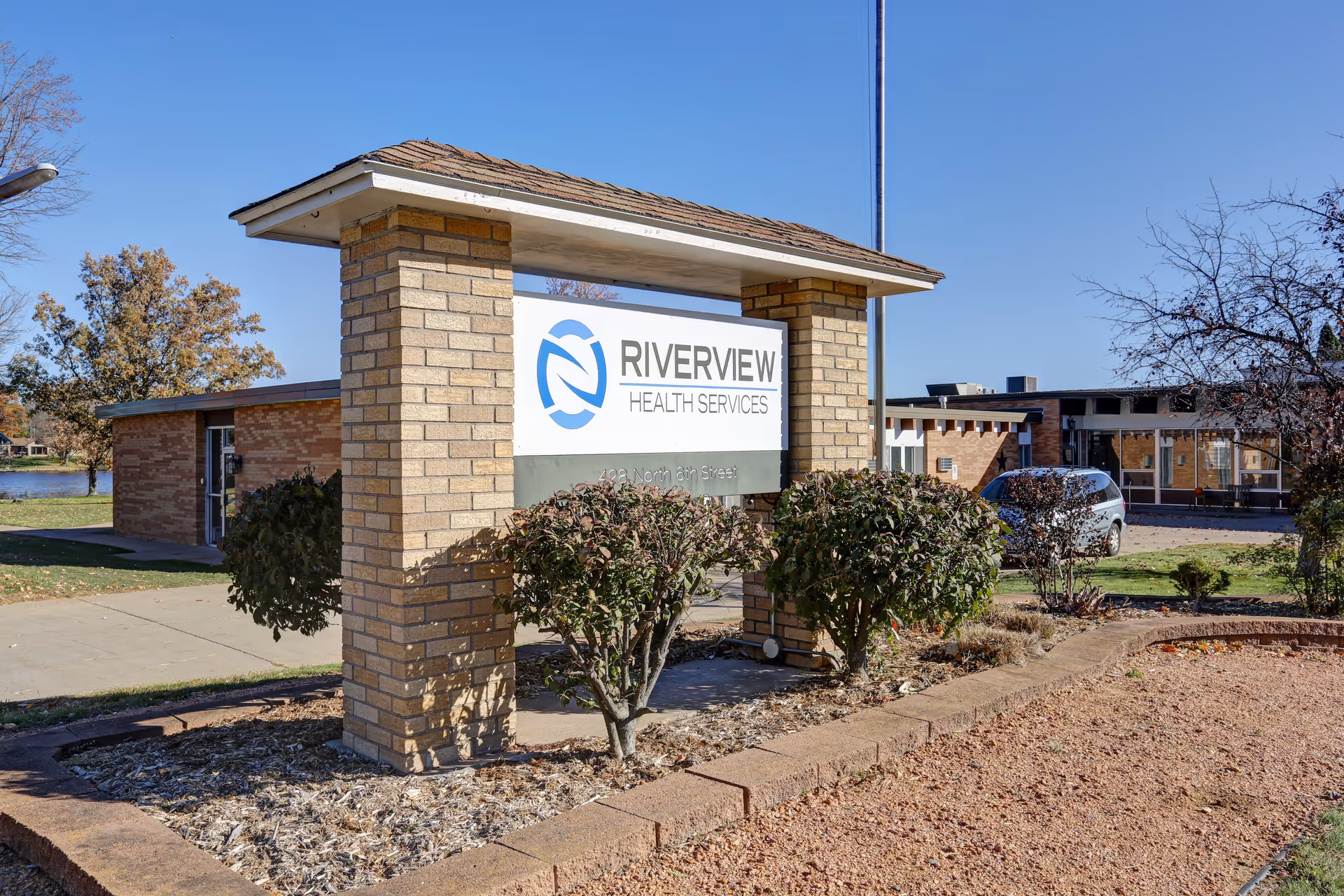 Outdoor sign reading "Riverview Health Services" in front of a brick facility building with shrubs and a parked car.