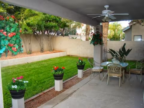 Covered patio area with a ceiling fan, glass-top table, and four chairs. The patio overlooks a small, well-maintained grassy yard bordered by a low wall and several plants and flowers in pots and garden beds.